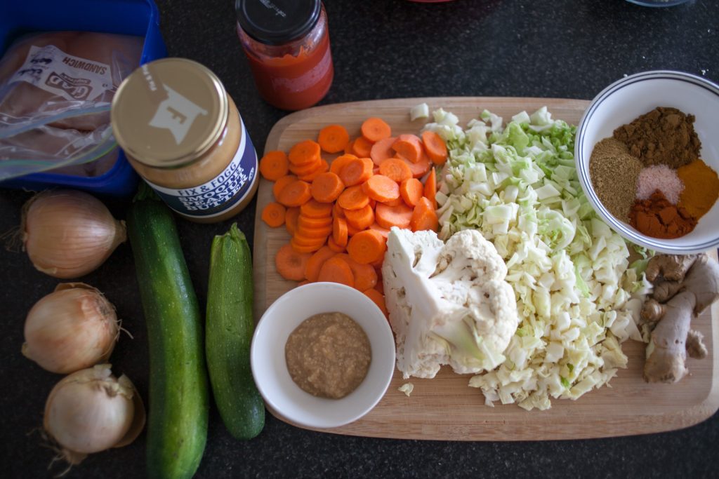 cutting board with raw, chopped veggies, peanut butter, ketchup, raw chicken, and a bowl of spices