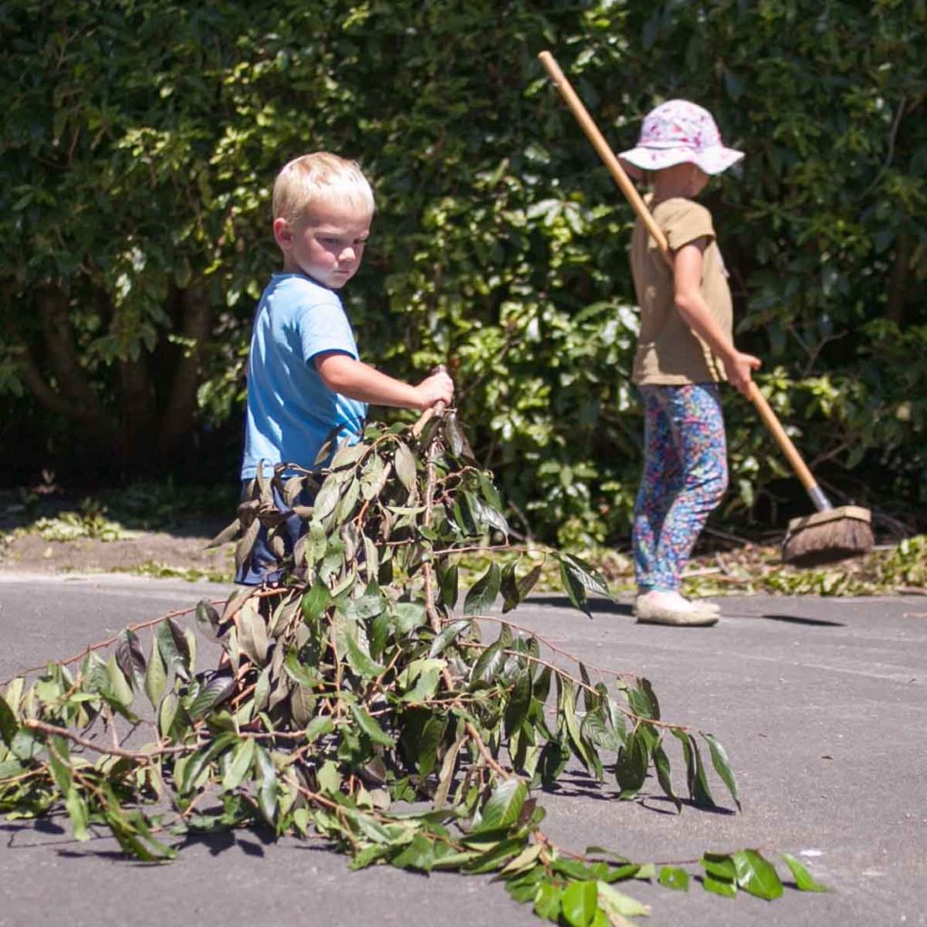 Little boy pulling branches over driveway while little girl sweeps in the background