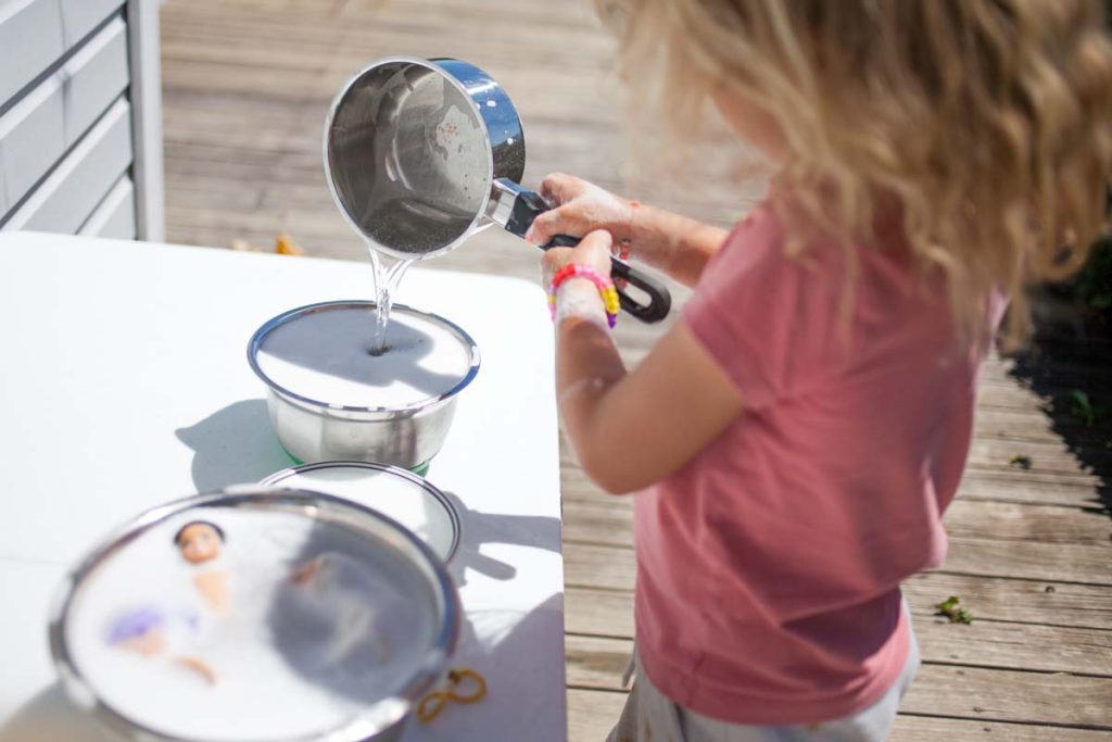 little girl pouring water from pot into mixing bowl on outdoor table