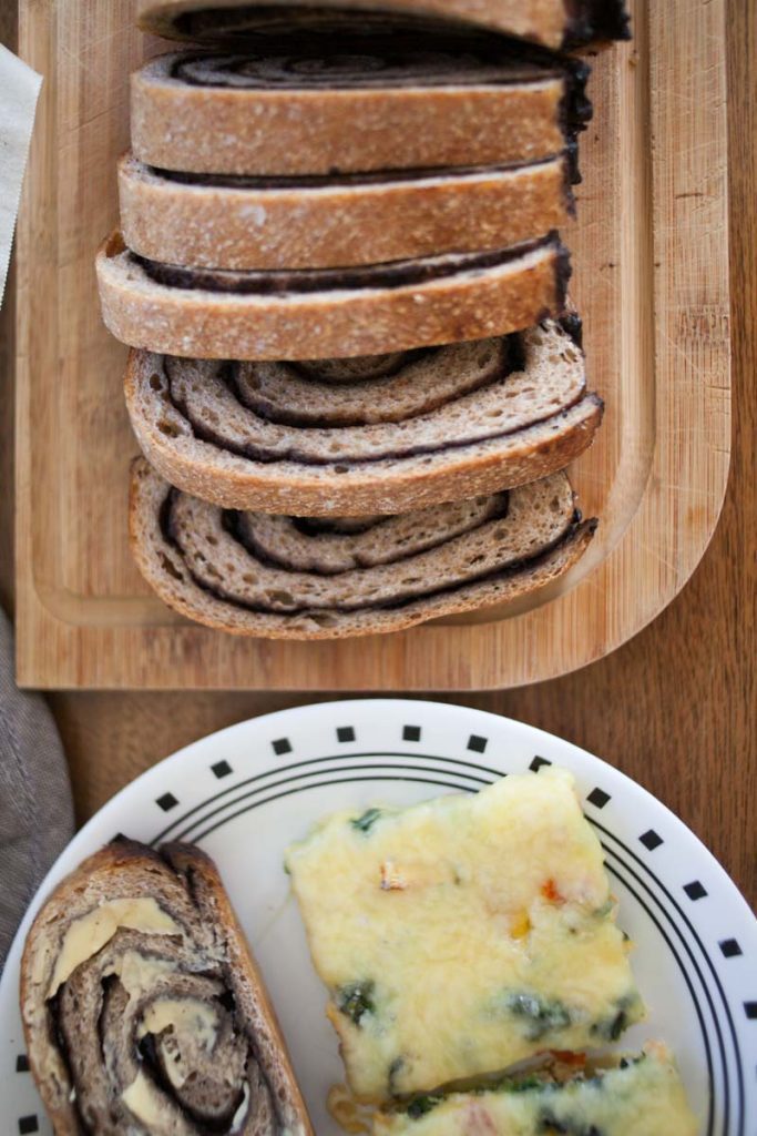 chocolate chai swirl bread slices on bread loaf next to plate with sheet pan scrambled eggs and slice of sourdough chocolate chai swirl bread