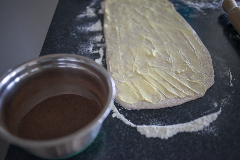 bread dough rolled out on counter with butter spread on top and bowl of chocolate chai sugar mix next to it.